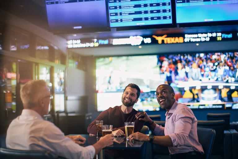 Three men enjoying drinks and conversation at a sports bar with multiple screens showing various games in the background.