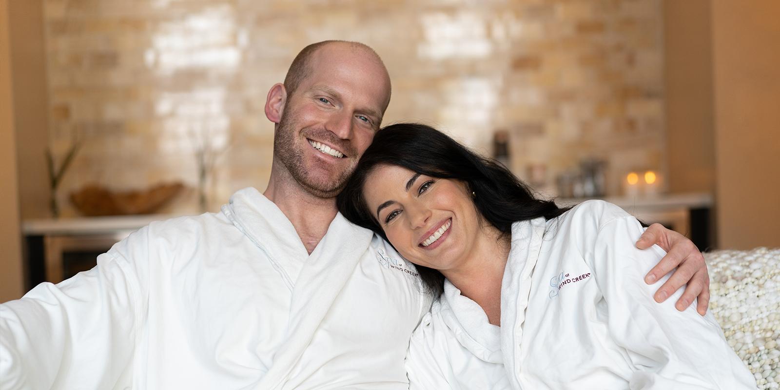 A smiling couple in a cozy, warm-toned spa room, both wearing white bathrobes and embracing each other