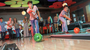 Two teen bows bowling while a group of teens watch and cheer