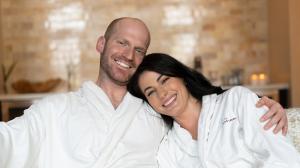A smiling couple in a cozy, warm-toned spa room, both wearing white bathrobes and embracing each other
